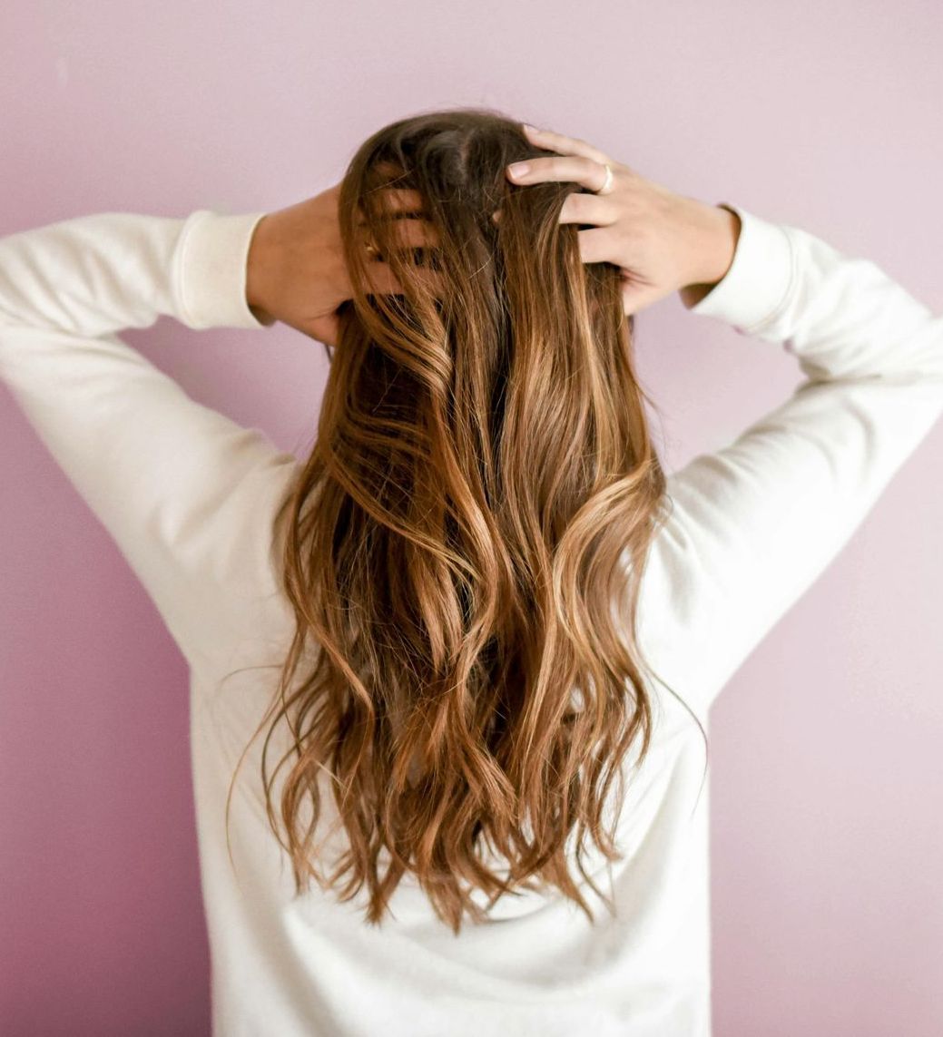 A woman in a white sweater is holding her hair in front of a pink wall.